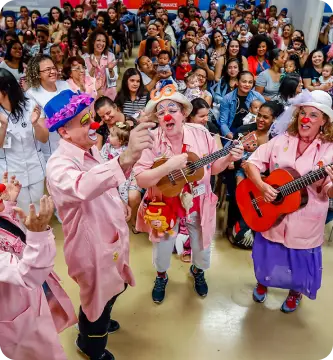 Grupo de voluntários do Einstein vestidos de palhaços tocando instrumentos e interagindo com pacientes e acompanhantes, representa o impacto positivo do voluntariado na humanização e acolhimento no ambiente hospitalar.