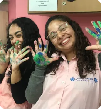 Voluntárias do Einstein sorrindo enquanto mostram mãos pintadas, simboliza a alegria e o impacto positivo do voluntariado, que promove acolhimento e humanização no ambiente hospitalar.